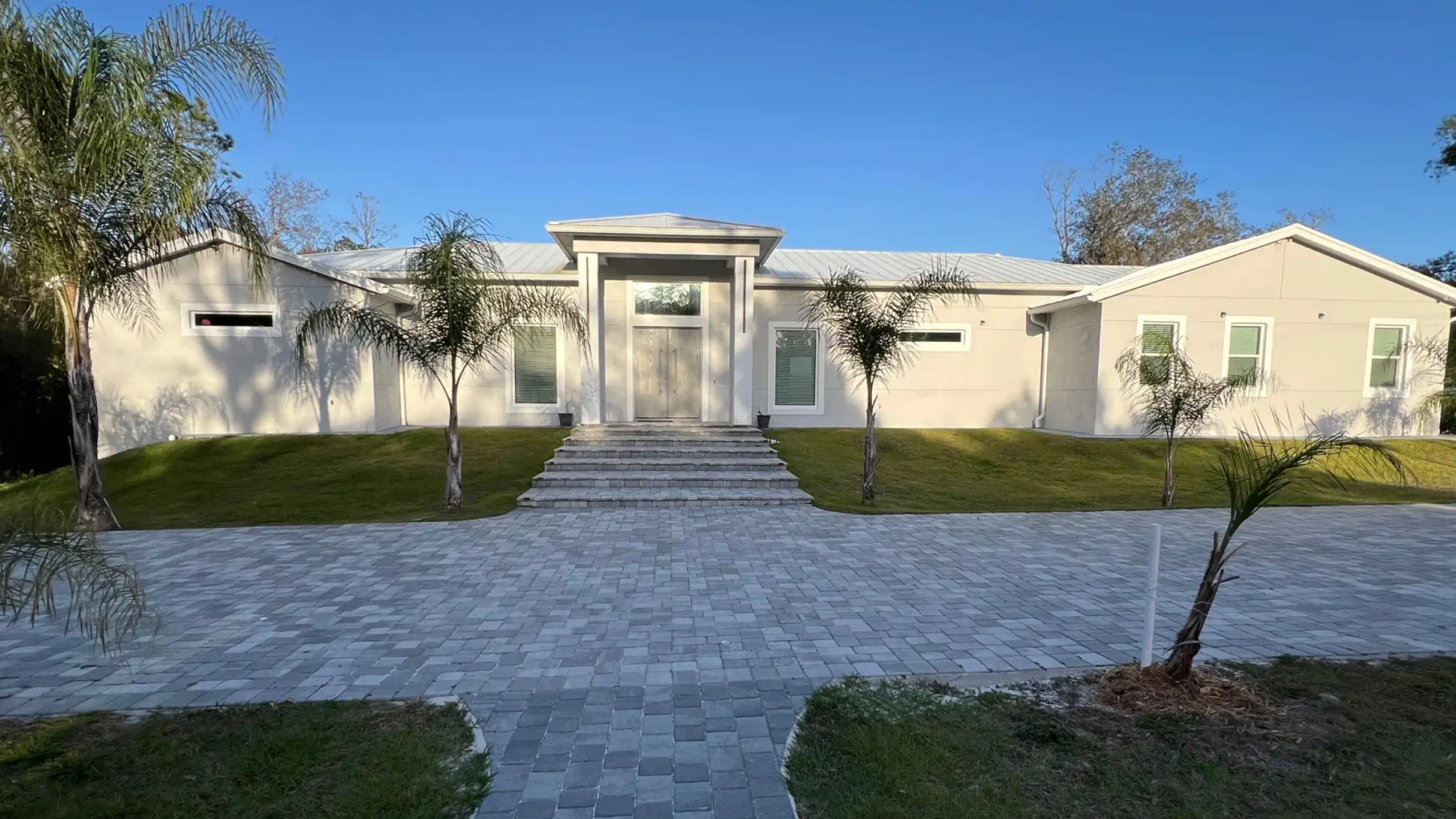light beige house with a light grey roofng some palms and a patio with grass around